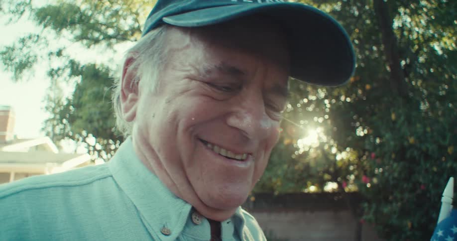 An elderly man smiles while gently tossing a ball and holding an american flag in his backyard