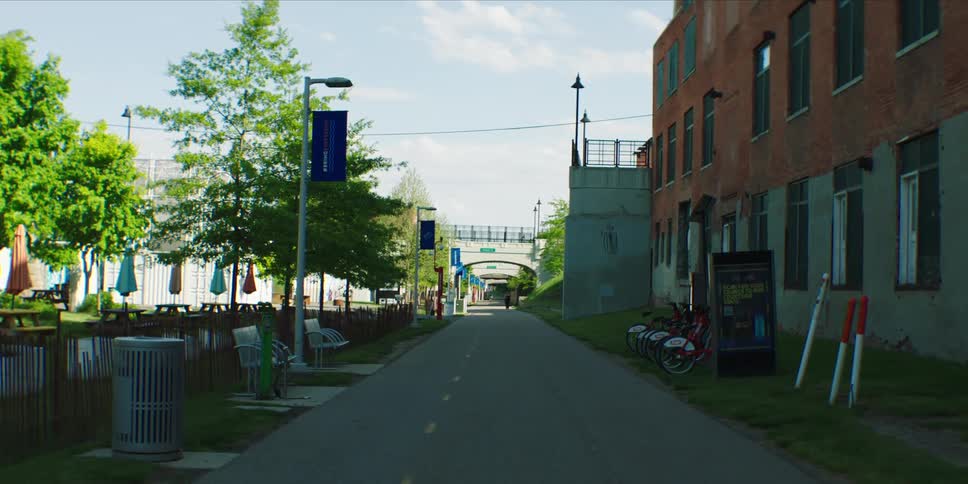 A quiet city bike path runs between a red brick building and a treelined park area with benches