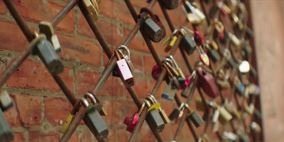 Dozens of colorful love locks are attached to a metal chainlink fence in front of a brick wall symbolizing everlasting love and commitment