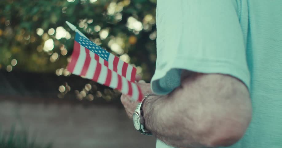 An older man walking near trees holding a small american flag