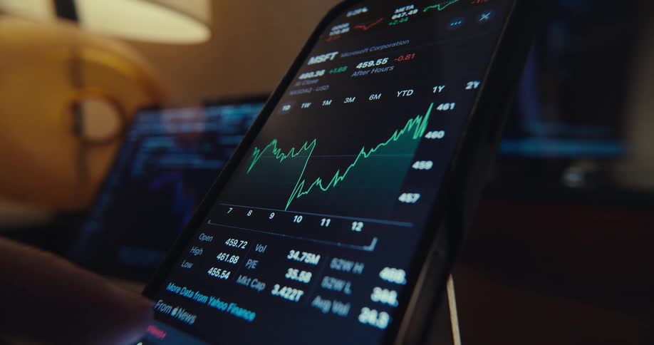A hand scrolls through stock market charts on a smartphone in a dimly lit room