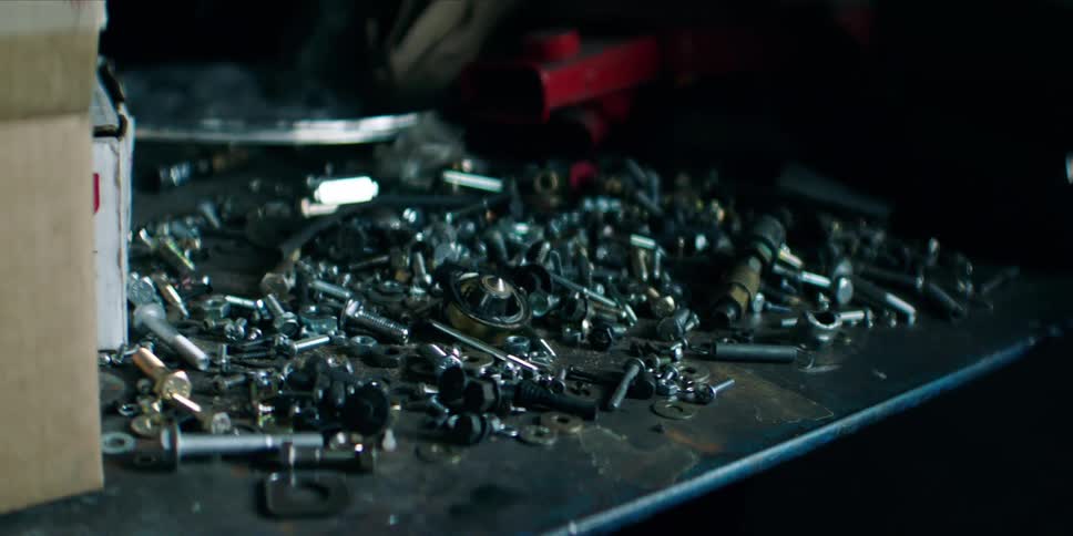 A messy pile of various nuts bolts and screws is scattered across a dark workbench in a garage or workshop