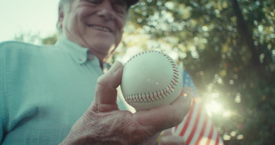 An elderly man smiling holding a baseball in one hand and an american flag in the other