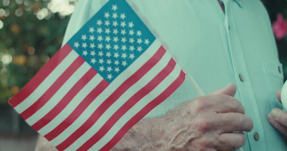 An elderly man holding a flag and a baseball while walking on a lawn at midday