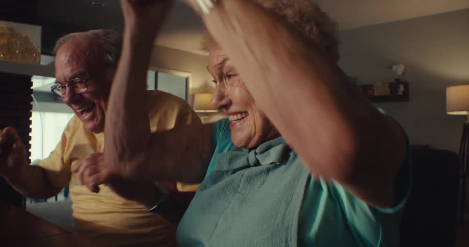 An elderly couple starts cheering while watching a tablet sitting on a desk in front of them