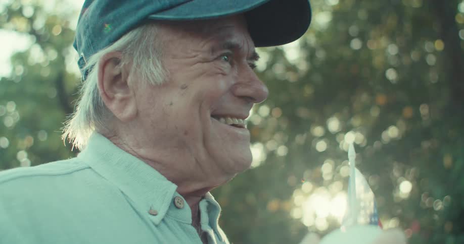 Elderly man smiles holding a baseball and flag in a sunny backyard