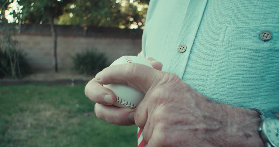 Elderly man smiles while standing and holding a baseball with a flag outdoors on a lawn during the day