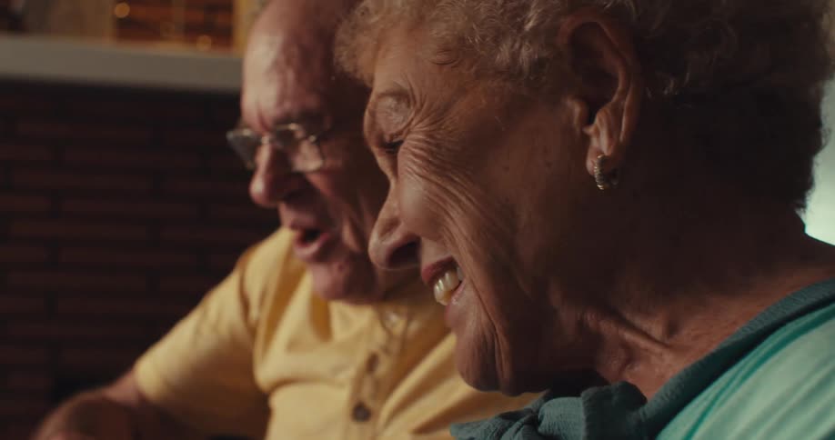 An elderly couple sit in a living room happily looking at something in front of them