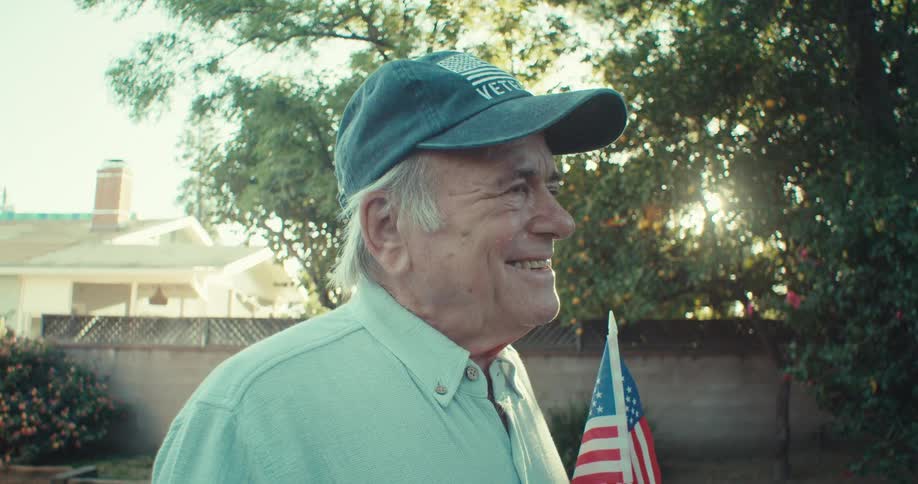 An elderly man stands on the lawn at midday laughing as he tosses a baseball into the air and catches it holding a small flag in his other hand