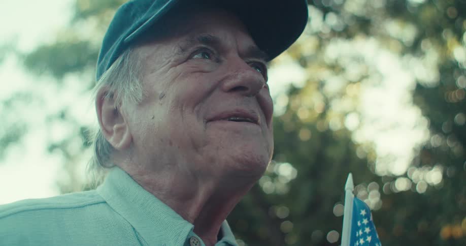 An elderly man smiling holding a baseball in one hand and a small american flag in the other