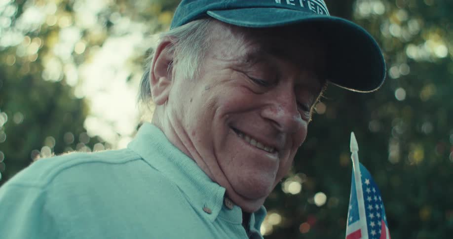 An elderly man in a cap smiles while holding a baseball and a small american flag in a sunny backyard
