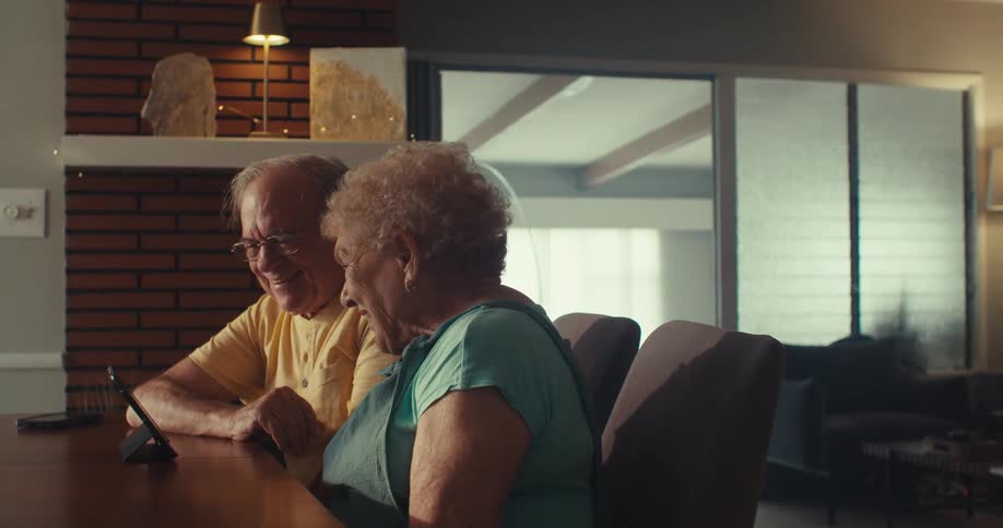 An elderly couple joyfully waves at a digital device during a video call in their cozy home