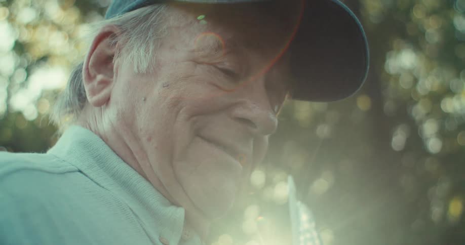 An elderly man outdoors near trees holding a baseball in one hand and a us flag in the other