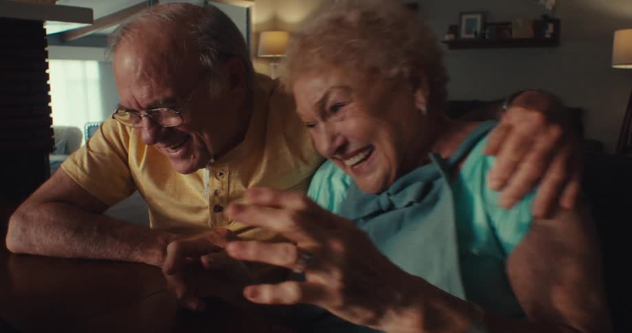 An elderly couple looking very happy while viewing a tablet placed on a desk indoors