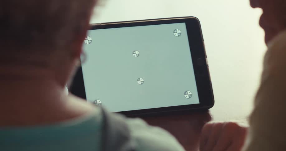 Two older adults sit closely while using a tablet with a green screen and tracking markers