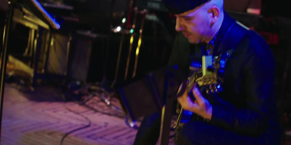 A man in a hat is holding a guitar and trying to press a button on a mixing console