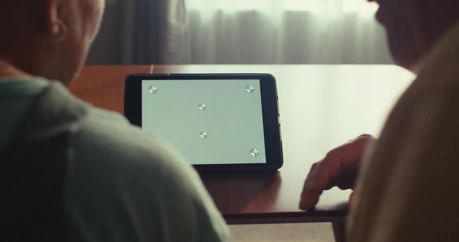 An elderly couple sits indoors while talking and looking at an electronic tablet
