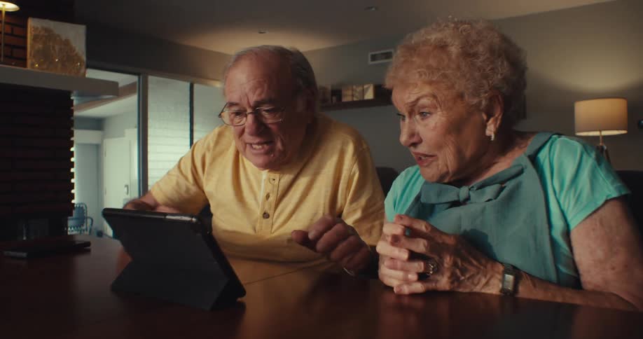 An elderly couple watching video on their tablet while at home become excited and happy