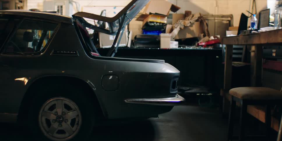 A parked vintage car with its trunk open inside a cluttered garage