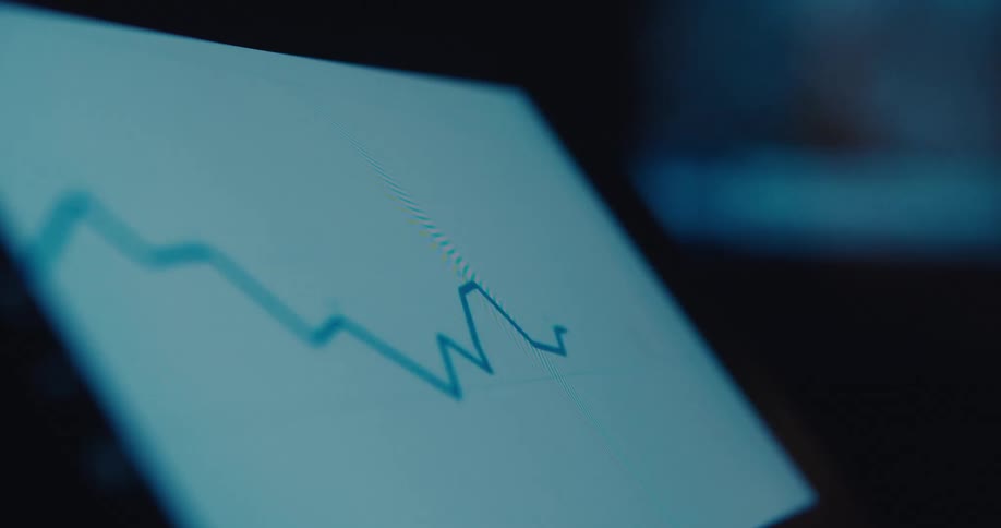 A downwardtrending blurry line graph is displayed on a tilted screen in a dimly lit setting