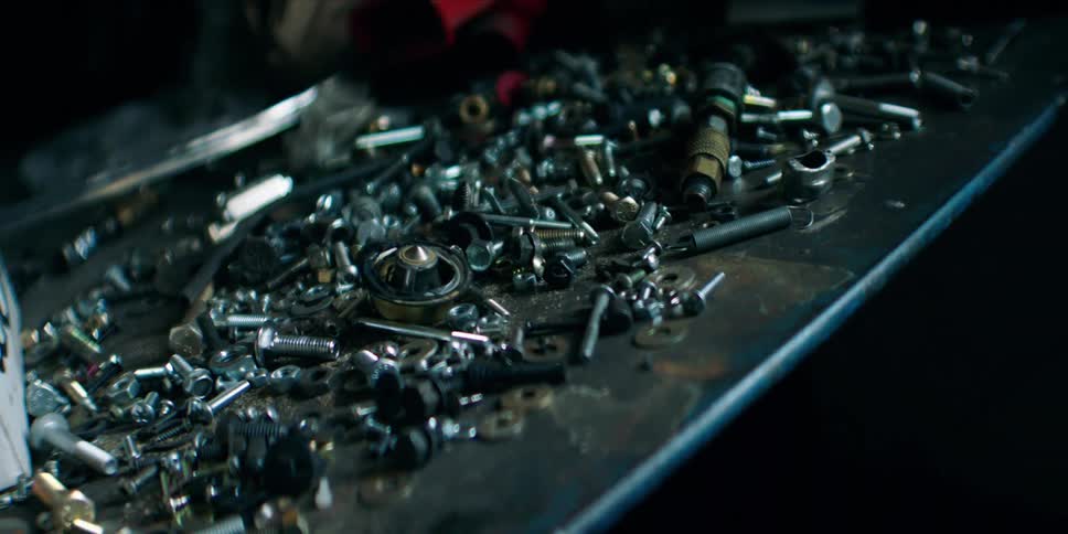A messy collection of various screws nuts bolts and other hardware sits on a dark metal workbench in a workshop