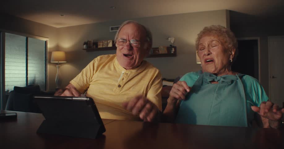 An elderly couple excitedly reacts while watching something on a tablet at their dining table