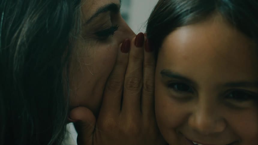 Mother and daughter whispers into each others ears while smiling in a room at daytime