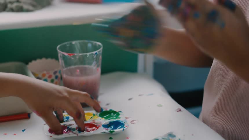 Two young childrens hands covered in colorful paint are pressing down onto a white surface with a palette of paints nearby