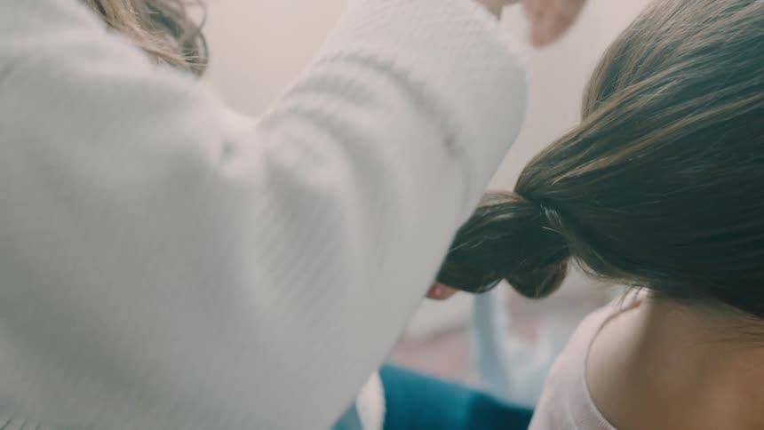 Mother ties daughters ponytail with a blue cloth while seated in a room