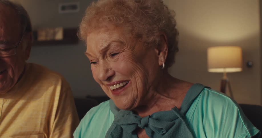 An elderly couple sits closely together smiling and looking at a tablet screen with joy