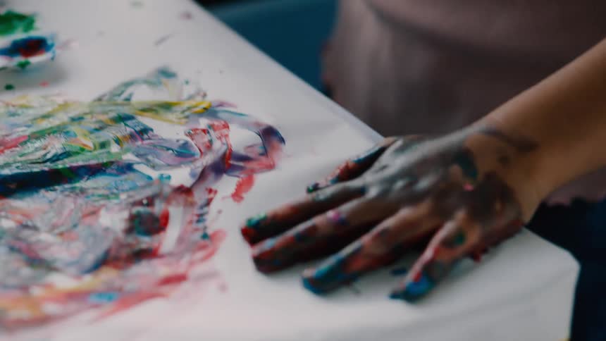 Childrens hands covered in paint press handprints onto a table with a watercolor palette nearby