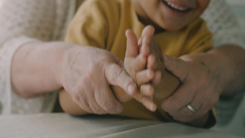 Elderly woman and a child clap together as she holds his hand from behind in a room