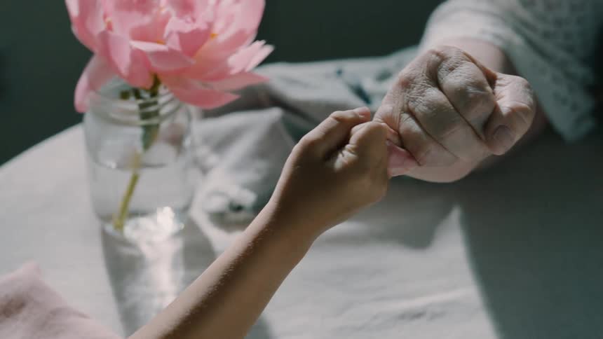 A child and an elderly person gently link pinky fingers across a softly lit table
