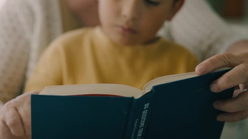 A young childs face is visible just above the open pages of a book as an adult helps him read