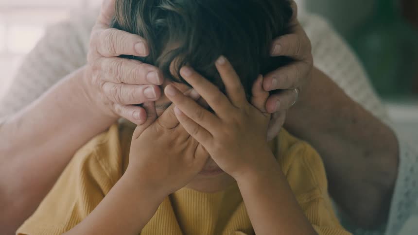 A small child with dark hair has their hands covering their face while an adults hands are gently placed around them