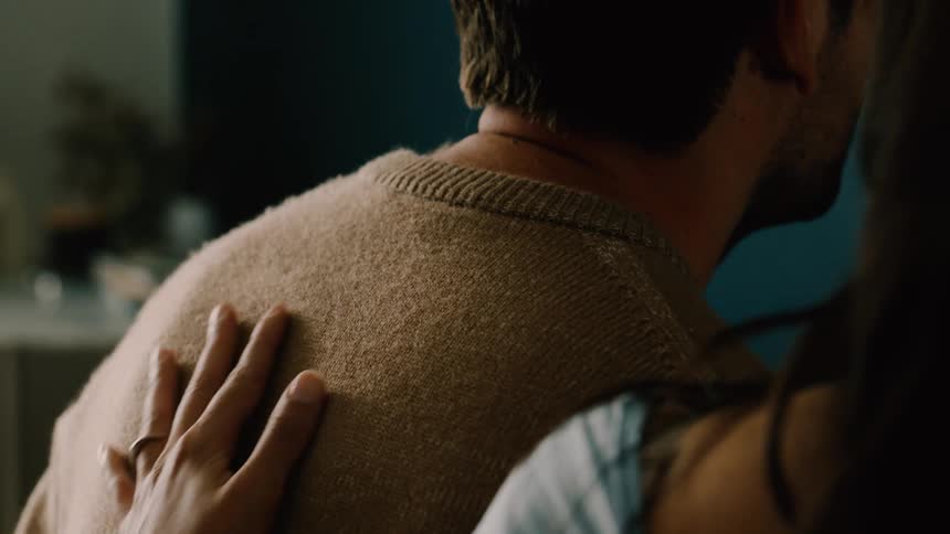 A woman gently embraces a mans neck and back in an intimate comforting moment indoors
