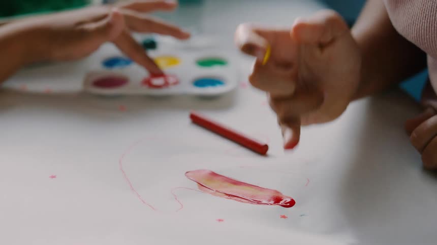 Two children dip their fingers in paint while making art on a white sheet