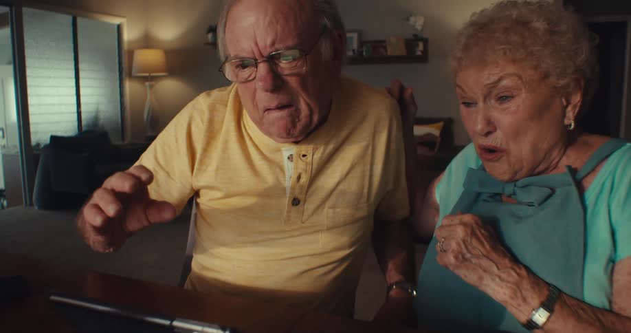 An elderly couple excitedly reacts while looking at a tablet screen together in a cozy living room