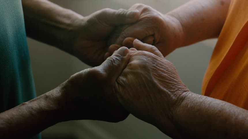 Two pairs of hands with wrinkled skin are gently touching inside a dimly lit room one wearing a ring