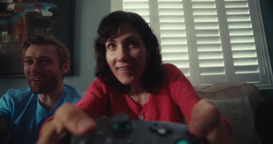 A smiling woman playing a video game while sitting on a couch with a man next to her and a vibrant abstract painting on the wall behind them