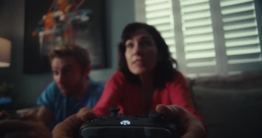 A man and a woman are excitedly playing video games on a couch in a living room with a large painting on the wall and window blinds