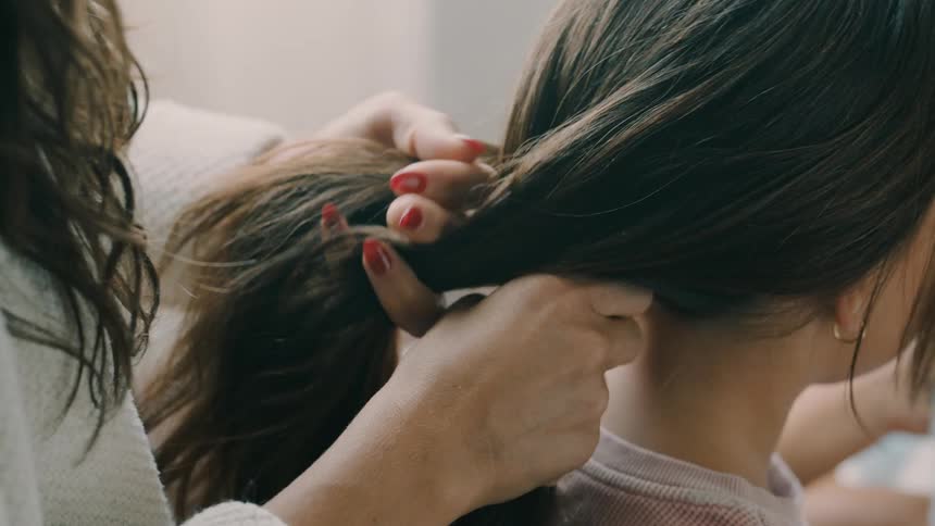 Closeup of a womans hands braiding a childs long dark brown hair in a room