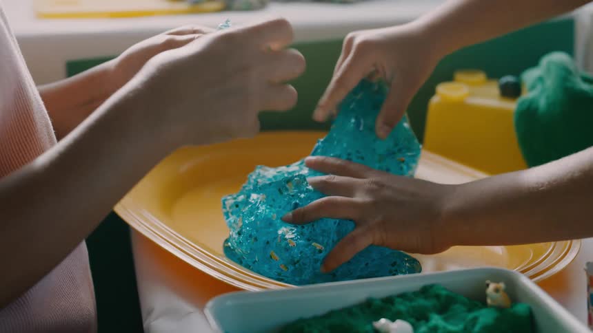 Two kids are indoors at midday molding blue slime on a yellow tray and cutting chunks off it as they play together