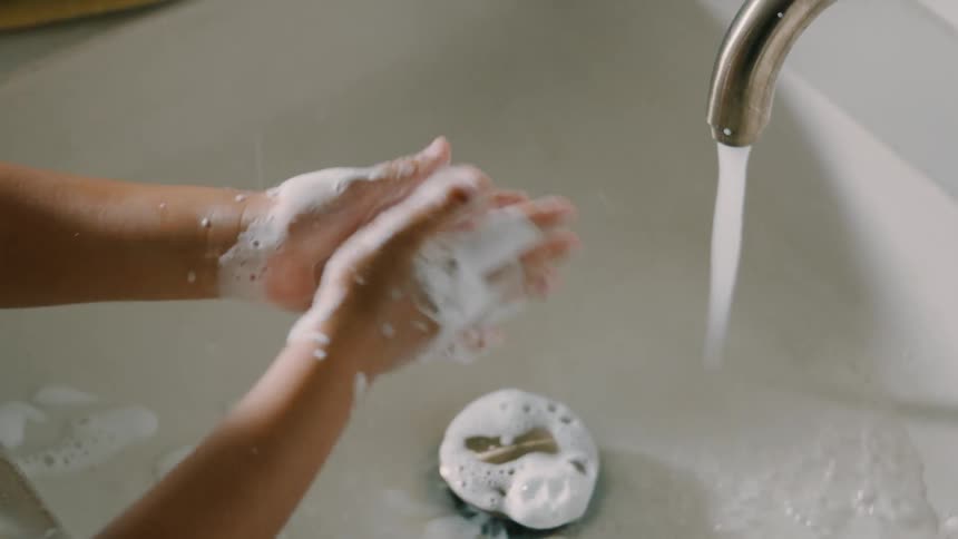 A childs hands covered in soap on top of a white sink with the tap running and pouring water