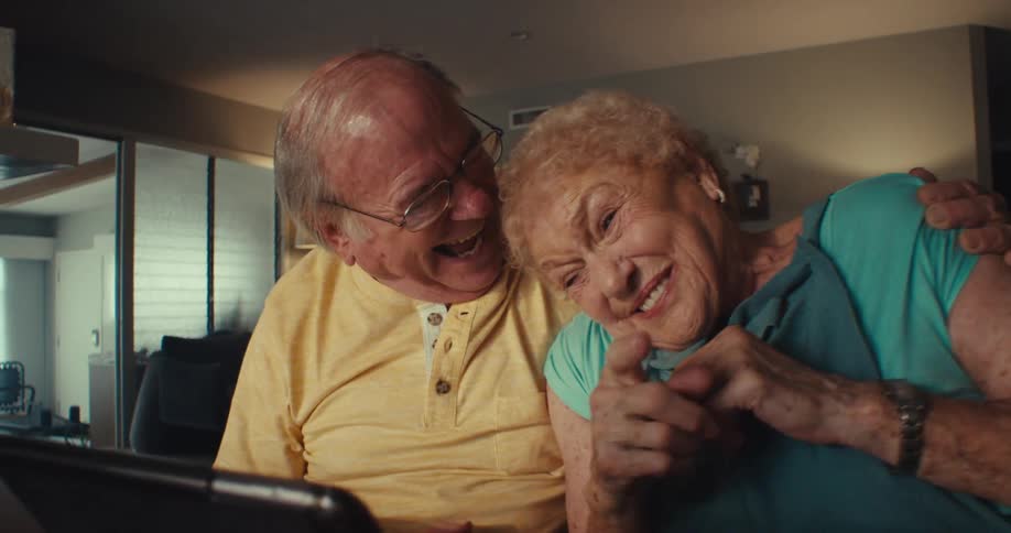 An elderly couple watching something on a tablet on the table the man hugs the woman and they look happy