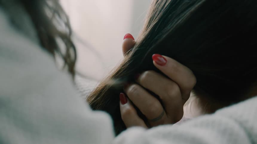 A pair of hands one with red manicured nails gently gathers and tends to a persons long dark hair against a light blurred background