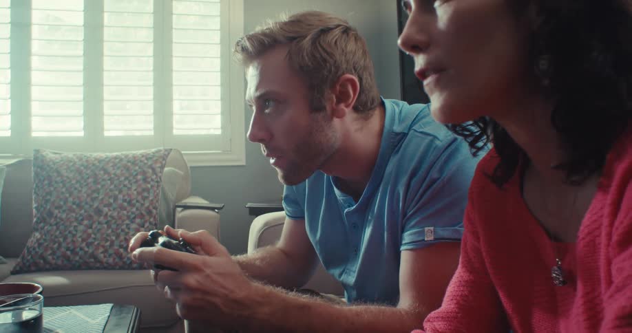 Man and woman play video games with controllers and converse while seated on a couch in a living room