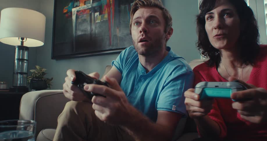 A couple sits on a couch playing a video game with great excitement at midday