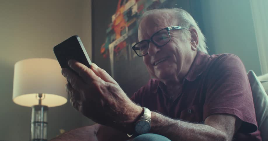 Cheerful elderly man sitting in living room and interacting with smartphone