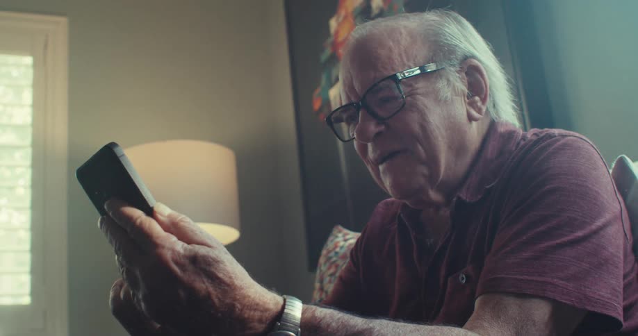 An elderly man wearing glasses is sitting happily while talking on the cell phone in his hand